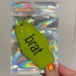 Hand holding a green keychain with 'brat' text against a reflective silver background