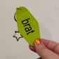 Hand holding a green keychain with 'brat' written on it against a plain background