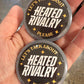 Two round metal objects with 'Heated Rivalry' text held in a hand.