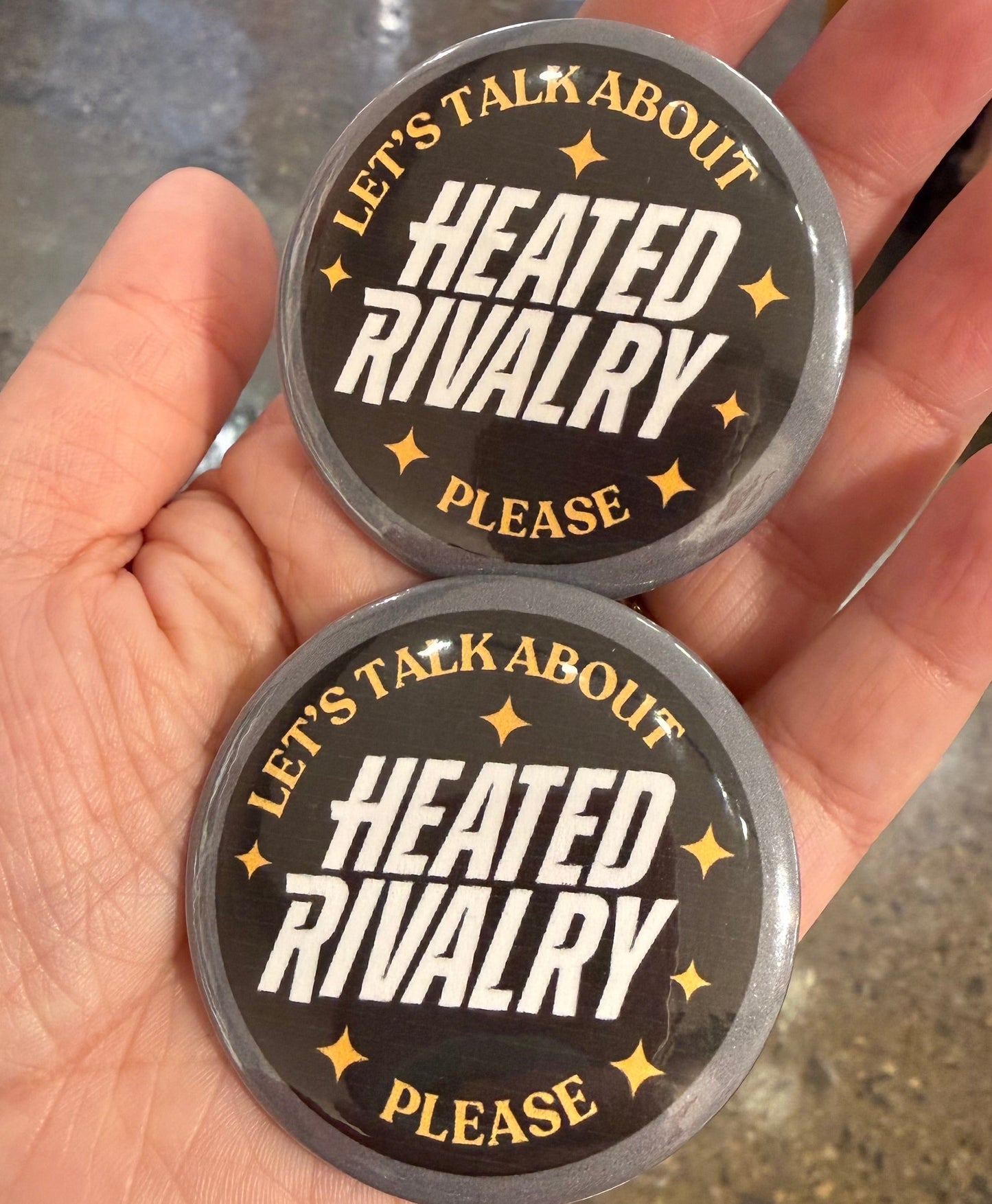 Two round metal objects with 'Heated Rivalry' text held in a hand.